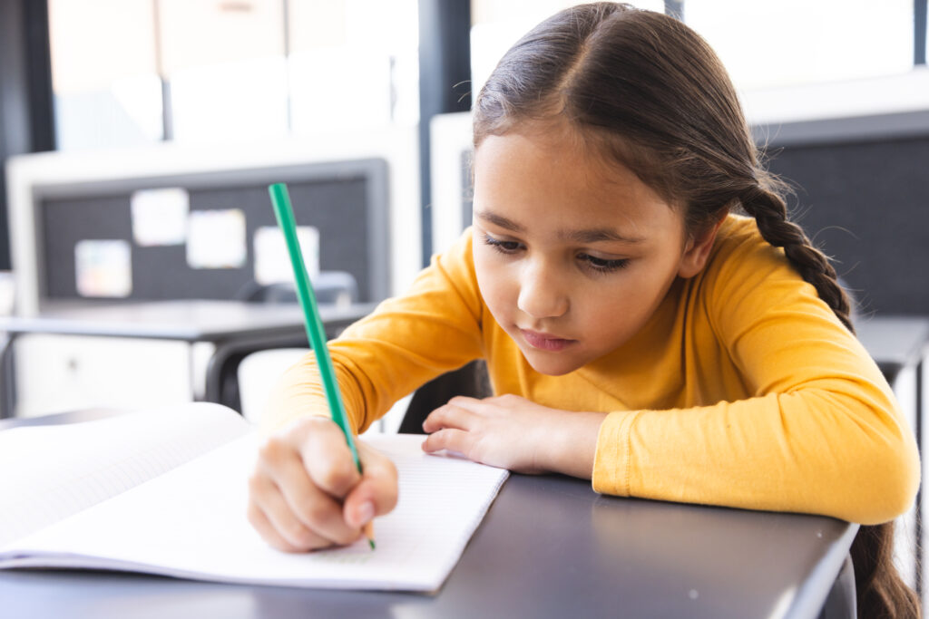 in school, young biracial girl with light brown skin focusing on writing in the classroom. she has long dark hair, wearing a yellow top, unaltered